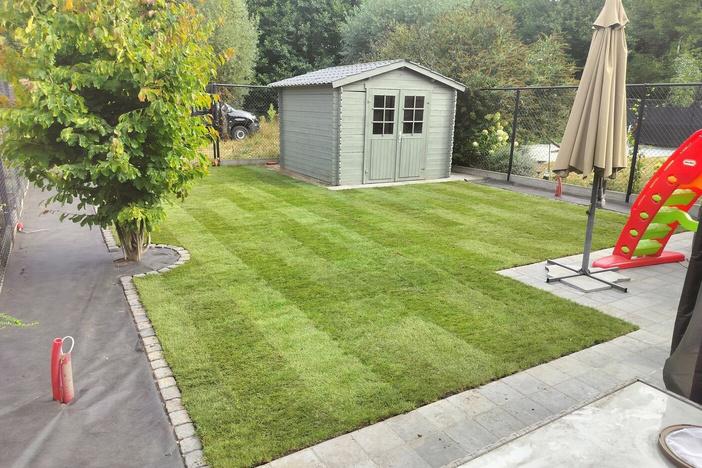 Dezelfde tuin met een nieuw gestreept gazon, een boordsteenrand en een jonge boom bij het tuinhuis